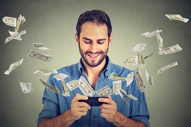 Technology online banking money transfer, e-commerce concept. Happy young man using smartphone with dollar bills flying away from screen isolated on gray wall office background